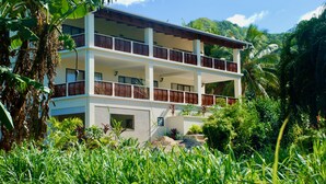 A two-story building with a balcony on each floor, surrounded by lush greenery and a mountain in the background.