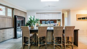 Una cocina moderna con una isla central, cuatro taburetes de mimbre, un refrigerador de acero inoxidable y una planta en maceta.