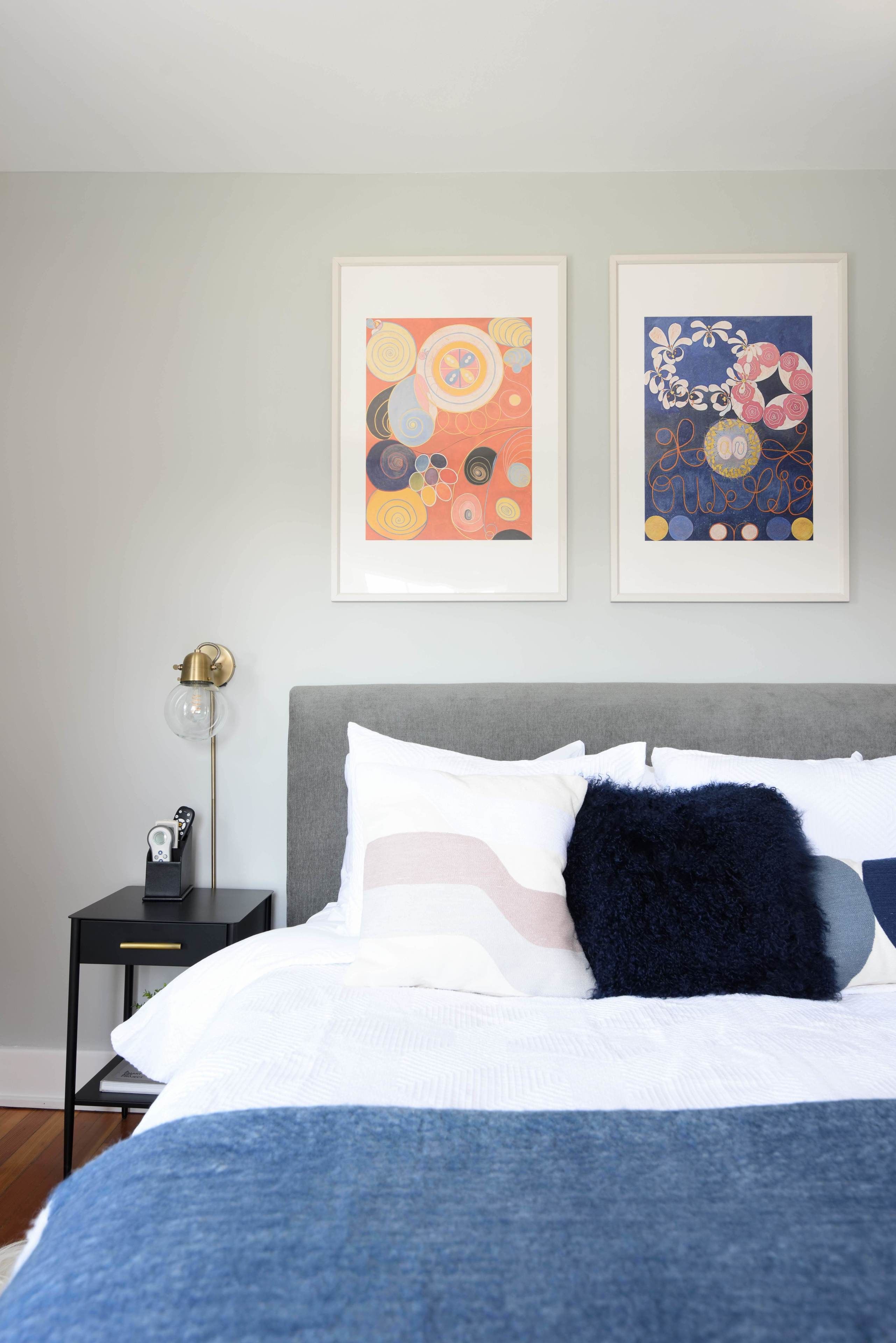 Un dormitorio con cama, almohadas, mesita de noche, lámpara y dos cuadros en la pared.