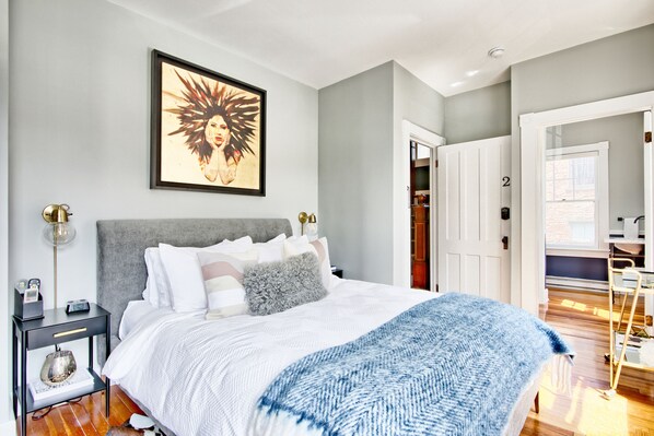 A bedroom with a bed, bedside table, lamp, and a framed artwork on the wall. - The Nest Hudson (Hudson)