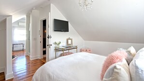A bedroom with a large bed, a desk with a mirror, a television on the wall, and a chandelier. - The Nest Hudson (Hudson)