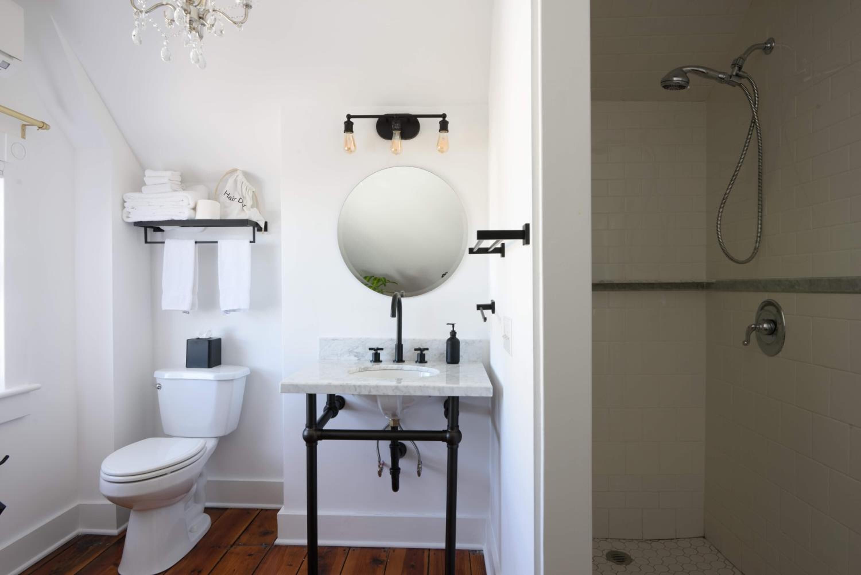Un baño con inodoro blanco, espejo redondo, lavamanos con tuberías negras, toallero y ducha.