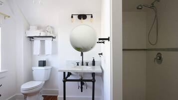 Un baño con inodoro blanco, espejo redondo, lavamanos con tuberías negras, toallero y ducha.