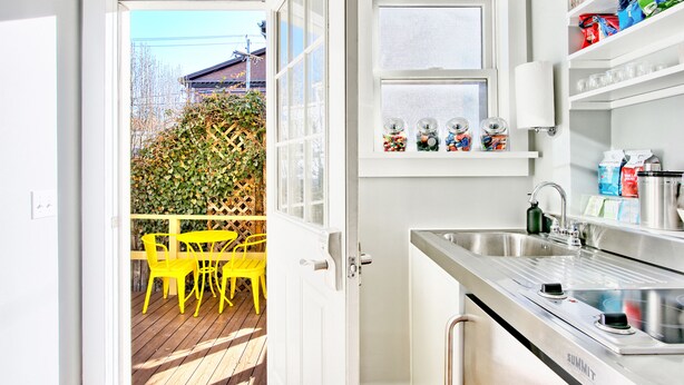 A kitchen with a stainless steel sink, a window with a view of a wooden deck and yellow chairs, and a door leading to an outdoor area. - The Nest Hudson (Hudson)