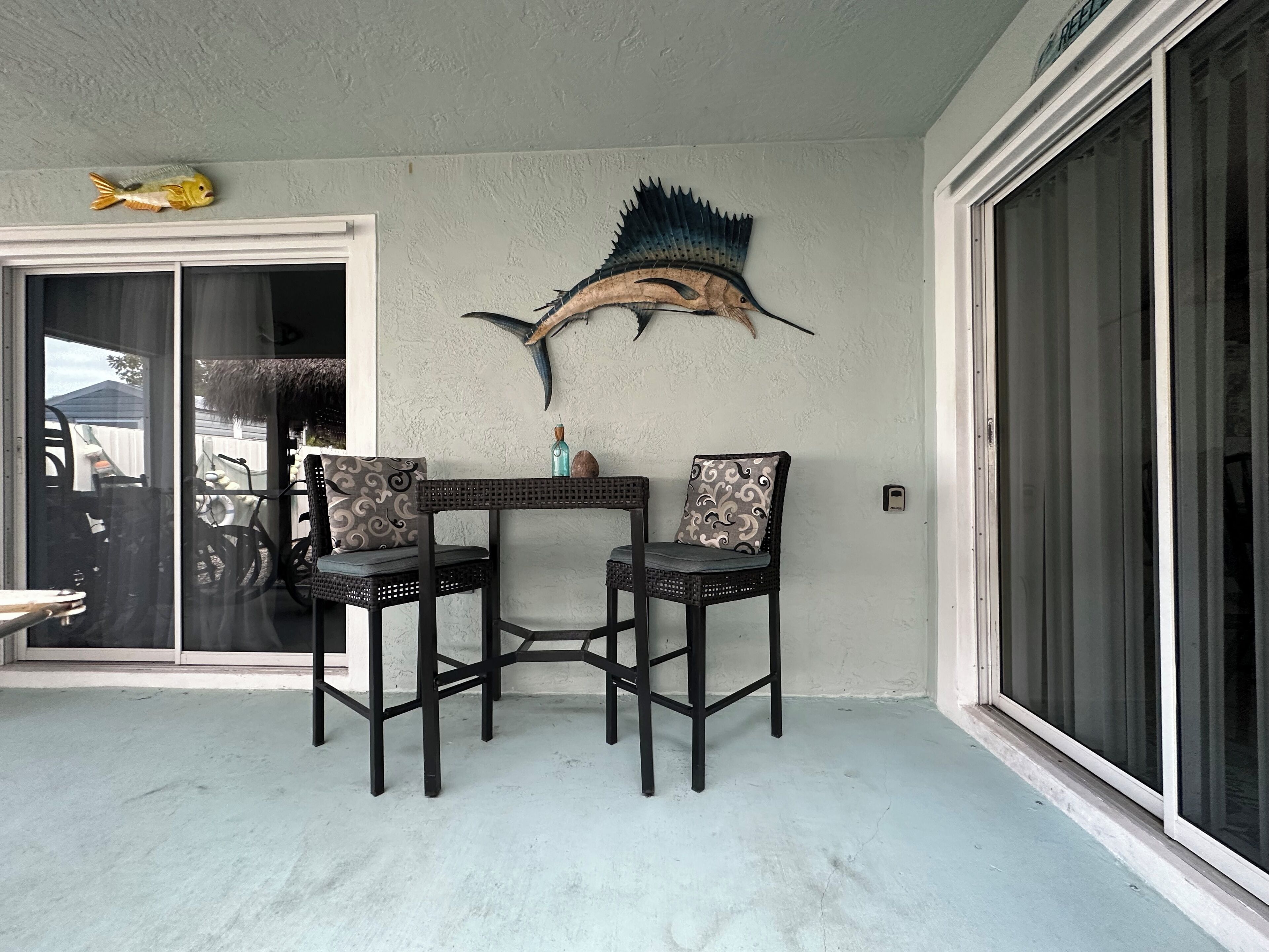 Un patio con dos sillas de mimbre, una mesita y peces decorativos de tiburón y pez colgados en la pared.