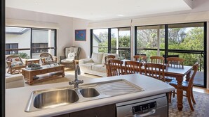 A spacious kitchen with a central island, a dining area with wooden chairs, and large windows offering a view of the outdoors. - Yabbarra Sands - Enjoy the beachside lifestyle- Close to Surf, MBT and Whales! (Dalmeny)