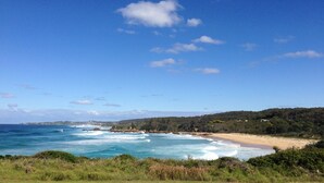 A coastal landscape with a beach, ocean waves, and a hilly background. - Yabbarra Sands - Enjoy the beachside lifestyle- Close to Surf, MBT and Whales! (Dalmeny)