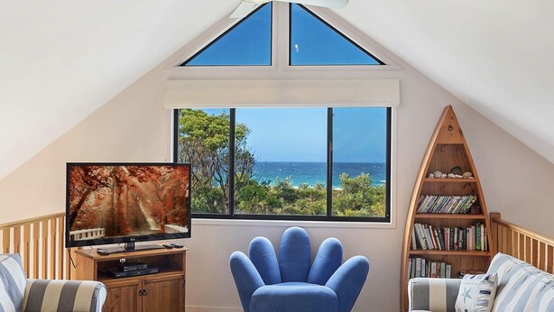 A cozy room with a TV, a blue chair, a bookshelf, and a view of the ocean. - Yabbarra Sands - Enjoy the beachside lifestyle- Close to Surf, MBT and Whales! (Dalmeny)