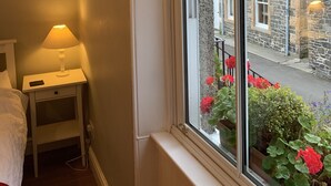 Una habitación con ventana a la calle, una mesita de noche con lámpara y una cama con cubrecama blanco.