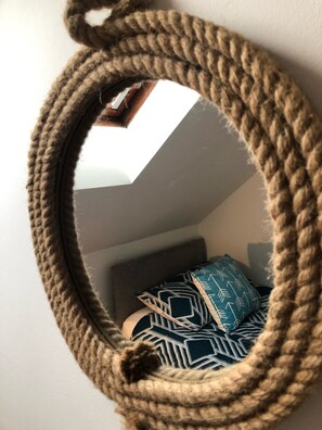 A round mirror with a rope frame, reflecting a bed with patterned pillows and a geometric wall design. - Large Traditional Stone 3 Bedroom 1ST Floor Apartment On The Jurassic Coast. (Langton Matravers)