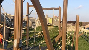 A playground structure with ropes and wooden beams, surrounded by grassy areas and stone buildings. - Large Traditional Stone 3 Bedroom 1ST Floor Apartment On The Jurassic Coast. (Langton Matravers)