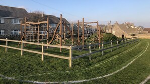 A playground with wooden structures and a grassy field, surrounded by stone houses. - Large Traditional Stone 3 Bedroom 1ST Floor Apartment On The Jurassic Coast. (Langton Matravers)