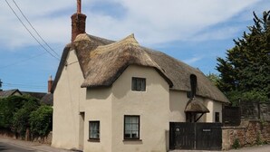 Una casa tradicional con techo de paja, una chimenea, un ventanal pequeño y un portón con el número 12.