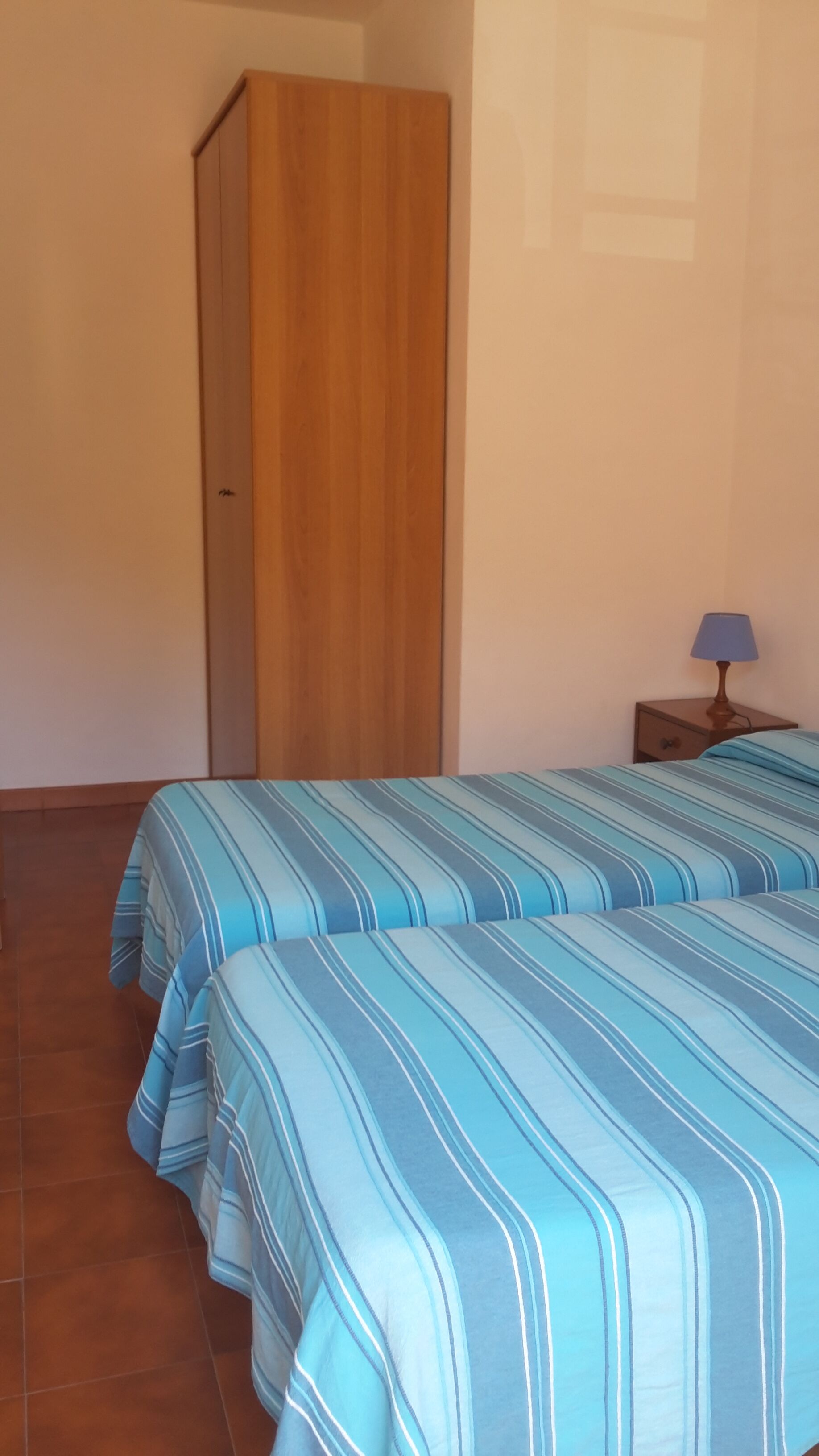 Ein Zimmer mit zwei Betten, einem Holzschrank, einem Nachttisch mit Lampe und einem gefliesten Boden.