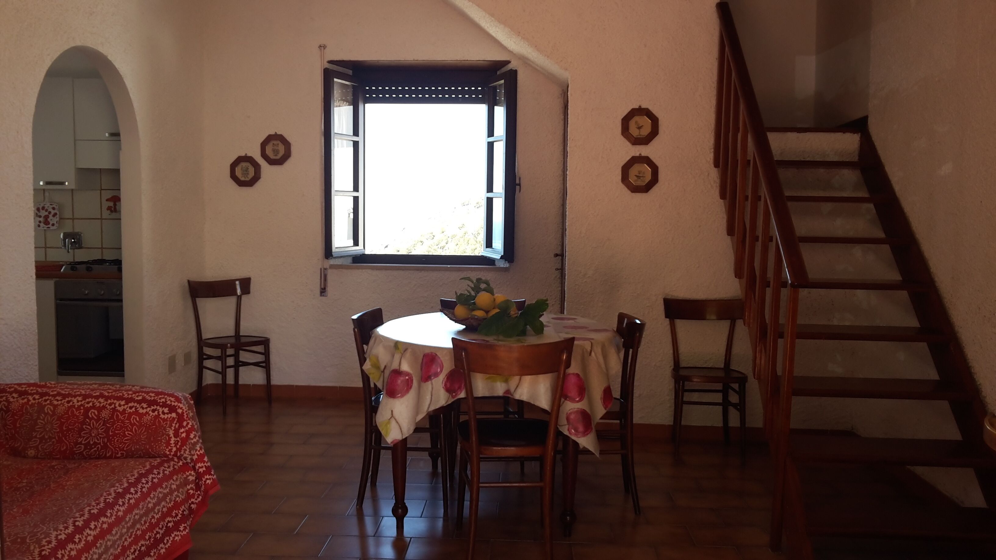 Un coin repas avec une table ronde, des chaises et une fenêtre avec des volets. Un escalier est visible sur la droite.