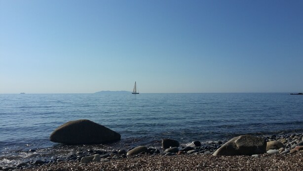 Una spiaggia ciottolosa con una barca a vela all'orizzonte e un masso in primo piano.