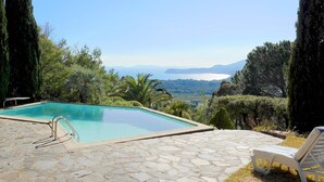 Una piscina con patio de piedra y una tumbona, rodeada de árboles y vistas a un lago y montañas.