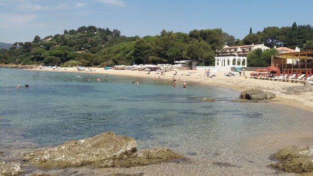 Una playa con agua cristalina, una orilla arenosa y árboles y edificios al fondo.