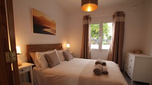 A neatly made bed with a headboard, a bedside table with a lamp, a nightstand, a dresser, and a window with curtains.