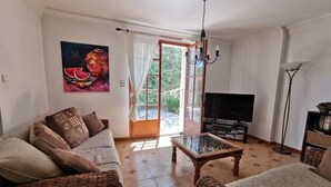 A living room with a sofa, a coffee table, a TV, and a floor lamp. There is a painting on the wall and a window with curtains. - Stylish villa, 13m private pool, tranquil garden, close to village amenities (Maureillas)