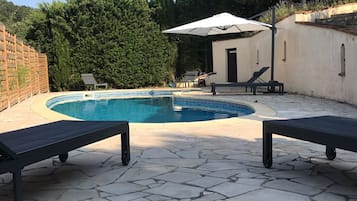 Une piscine avec deux chaises longues, un parasol et un petit bâtiment en arrière-plan.