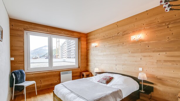 Een slaapkamer met houten lambrisering, een bed, twee nachtkastjes, een stoel en een groot raam met uitzicht op gebouwen en bergen.