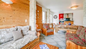 Een gezellige woonkamer met een houten bank, een eettafel en stoelen. Achtergrond toont een keuken met rode kastjes en een fornuis.