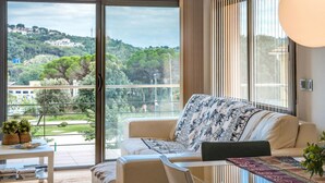A modern living room with a sofa, a table with books, and a view of a green landscape through large windows.
