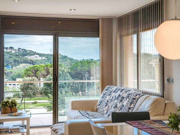 Un salon moderne avec un canapé, une table sur laquelle sont posés des livres et une vue sur un paysage verdoyant, visible à travers de grandes fenêtres.