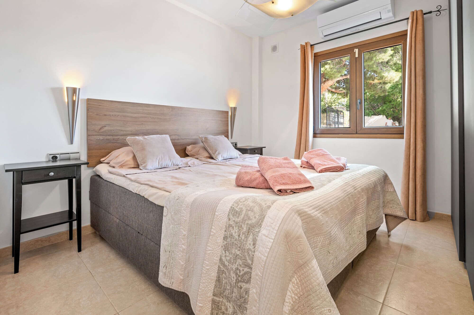 Un lit bien fait, recouvert d’une couvre-lit à motifs, deux tables de chevet avec des lampes, une fenêtre avec des rideaux et un appareil de climatisation fixé au mur.