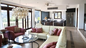 A modern living room with a white sofa, a glass coffee table, and a red armchair. A kitchen with stainless steel appliances is visible in the background. - Luxury modern villa with spectacular views of sea and heated private pool (La Herradura)