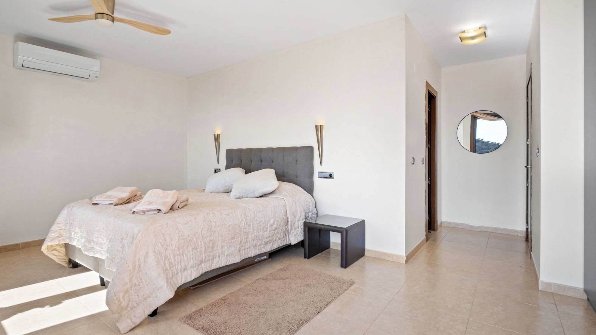 A bedroom with a bed, bedside table, wall-mounted lights, an air conditioner, and a mirror on the wall.