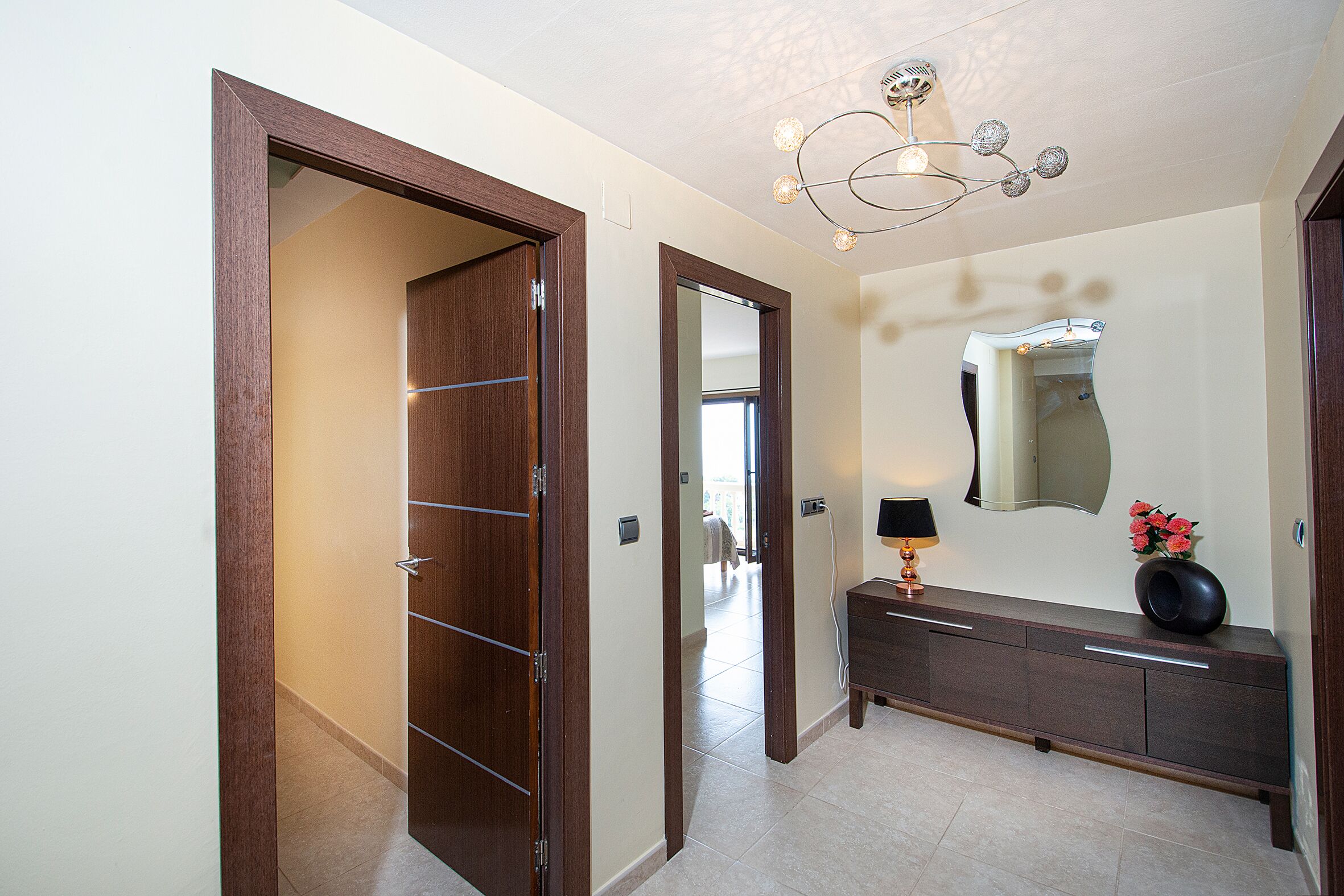 A hallway with a wooden door, a mirror, and a dark-colored console table with a lamp and flowers.