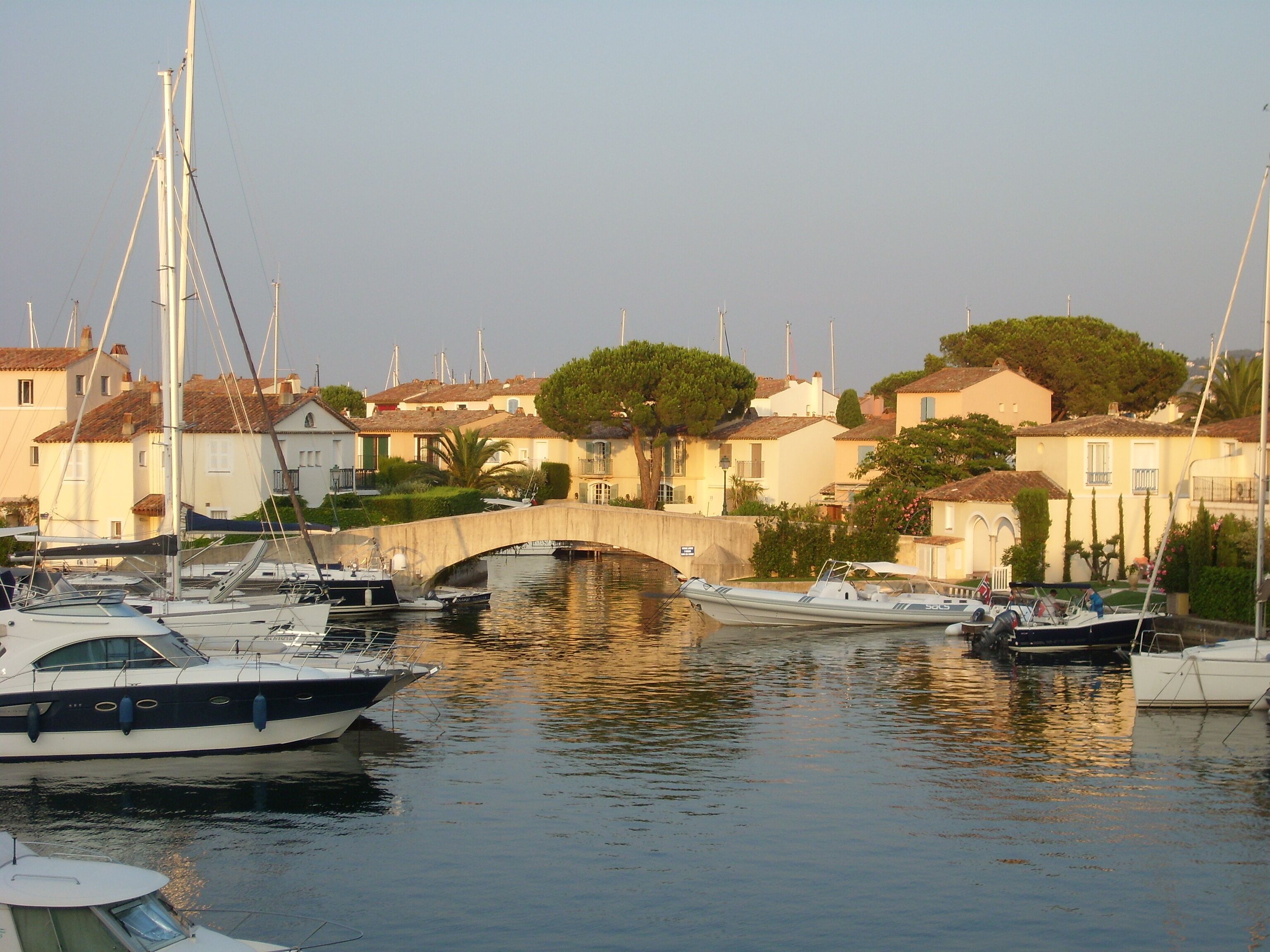 Un puerto deportivo con barcos amarrados, un puente de piedra y edificios de arquitectura mediterránea.