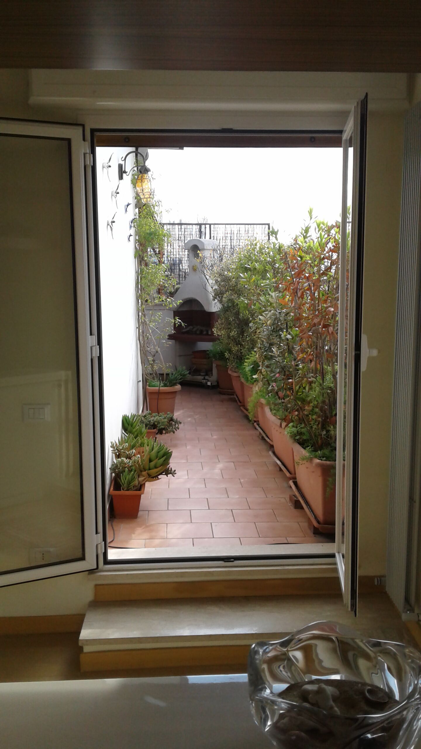 Una puerta corredera abierta que da a un patio con baldosas, plantas en macetas y una parrilla.