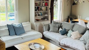 Een knusse woonkamer met een witte bank, een grijze bank en een houten tafel met een dienblad. Er is een boekenplank vol boeken en een hanglamp.
