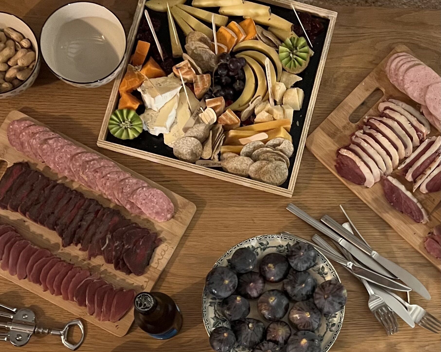 Forskellige madvarer, herunder skiveskåret kød, oste, frugt og nødder, anrettet på et træbord.
