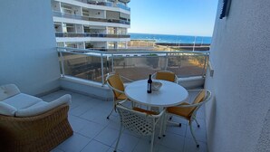 Un balcon avec une table et des chaises blanches, un canapé en osier et une vue sur la mer et les bâtiments.