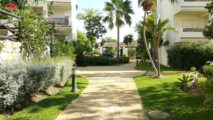 Ein gepflasterter Weg führt zu einem Gebäude mit Balkonen, umgeben von üppiger Vegetation und Palmen.
