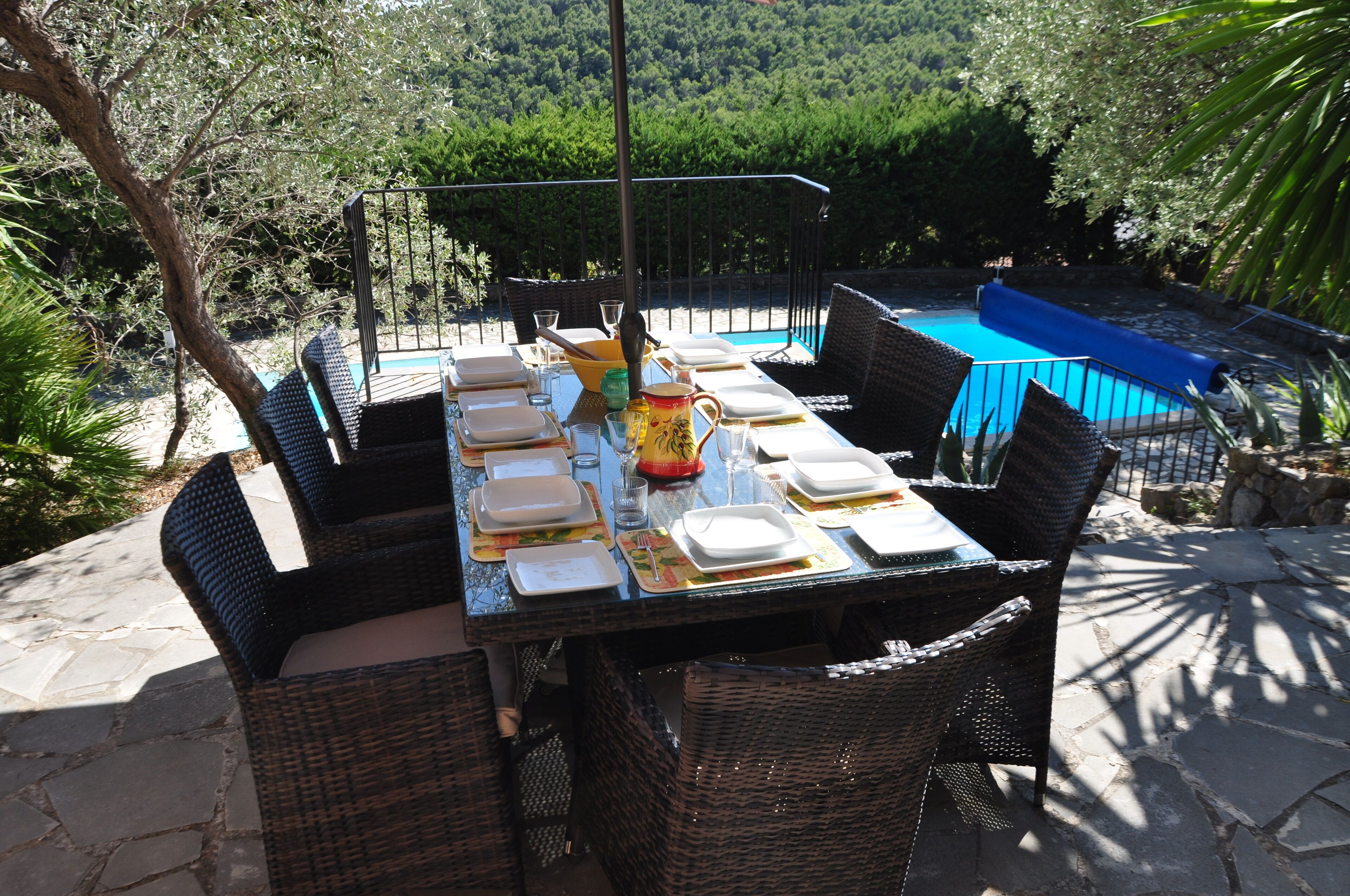 Un área de comedor al aire libre con una mesa preparada para cuatro personas, rodeada de sillas de mimbre, cerca de una piscina.
