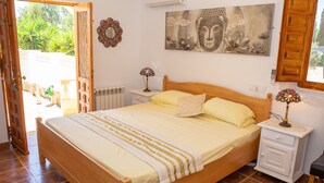 Een slaapkamer met een houten bed, een nachtkastje met een lamp, een wandairconditioner en een raam met uitzicht op groen.