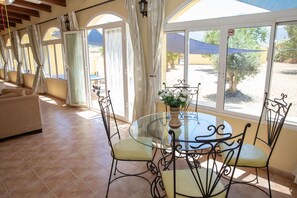 A room with a glass table, four chairs, a sofa, and large windows with curtains.