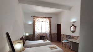 A room with two beds, a table, a chair, a lamp, and a window with curtains. - Town House Within Lagos Castle Walls (Lagos)