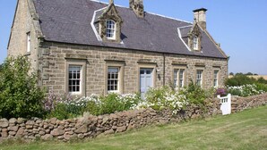 A stone building with a slate roof, multiple chimneys, and a stone wall with flowers. - Beautiful Stone Cottage - A Unique Rural Retreat In The Scottish Borders (Duns)
