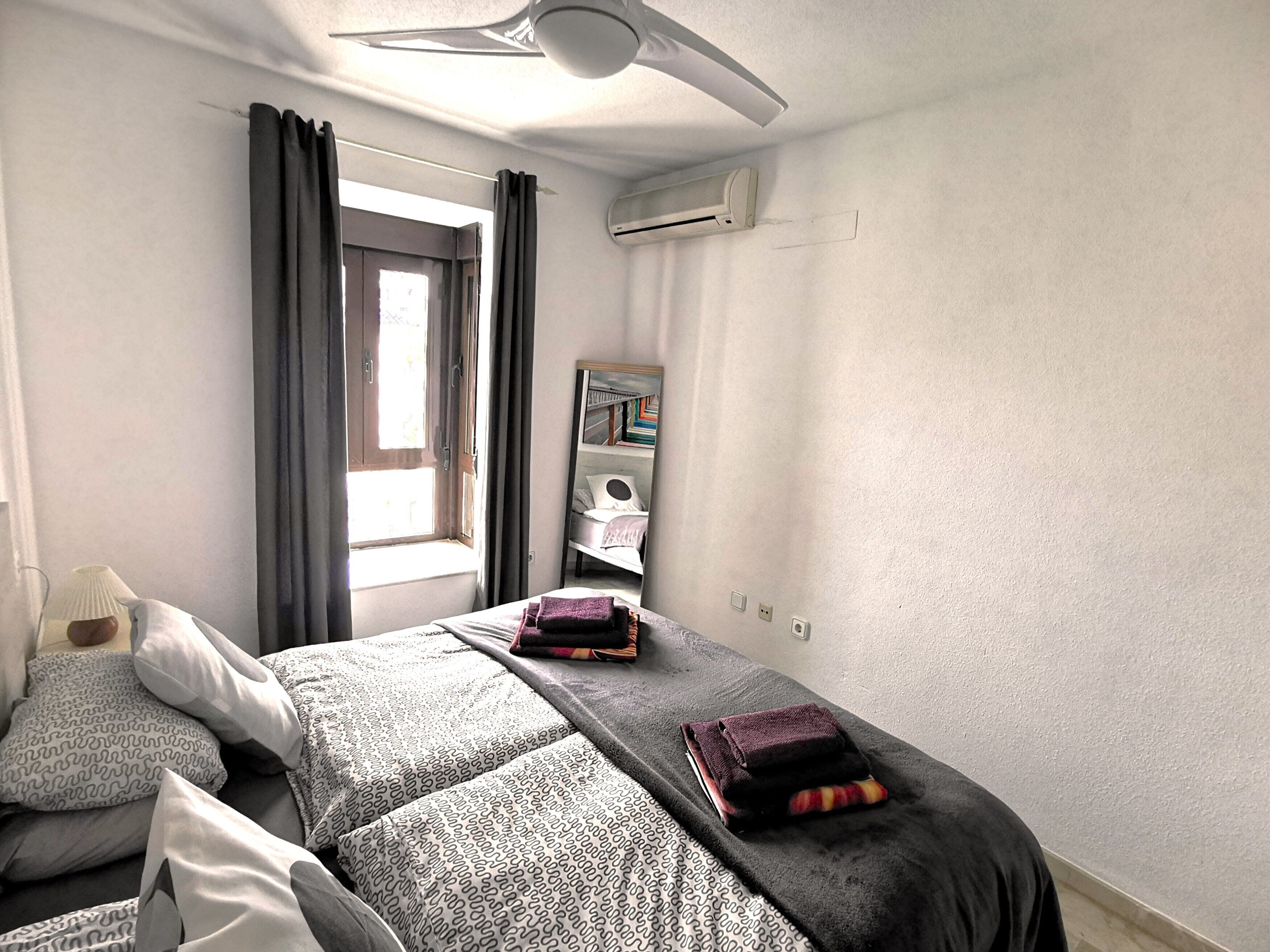 A hotel room with a bed, a window with curtains, a ceiling fan, an air conditioning unit, and a mirror on the wall.