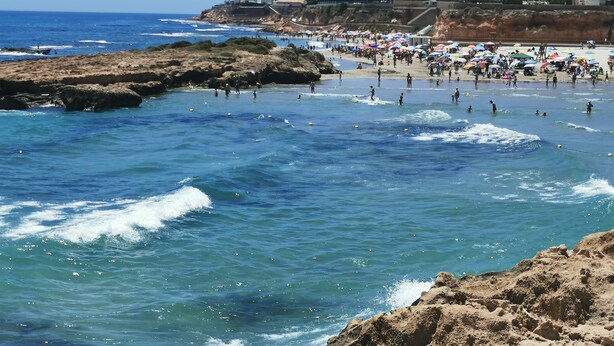 Une plage aux rivages rocheux et aux vagues déferlantes, une zone de plage très fréquentée avec des parasols, et des bâtiments en arrière-plan.