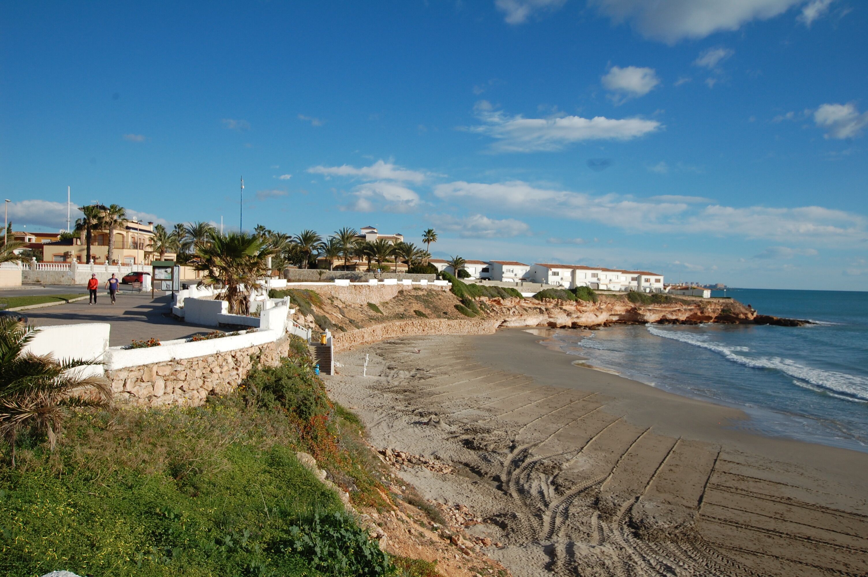 A coastal area with a sandy beach, a stone wall, and white buildings with red roofs.