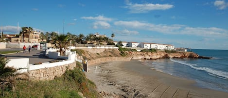 A coastal area with a sandy beach, a stone wall, and white buildings with red roofs.