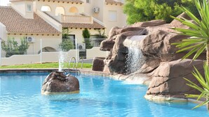 Une piscine agrémentée d’un élément rocheux et d’une cascade, entourée d’une clôture et bordée d’un bâtiment dont le toit est recouvert de tuiles.