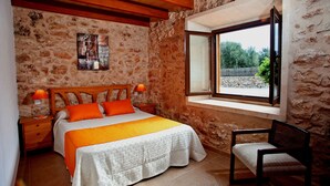 A bedroom with a stone wall, wooden ceiling beams, a large bed with orange bedding, a nightstand with lamps, and a window with a view of greenery.
